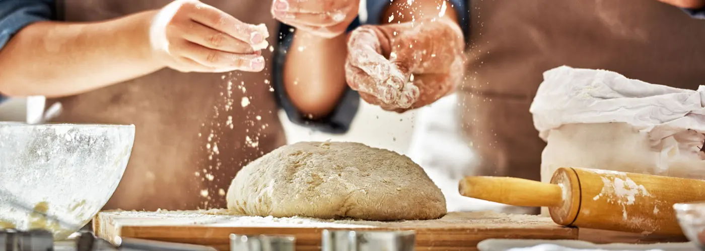 Man and Woman in a kitchen sprinkling flour over dough on a butcher block.