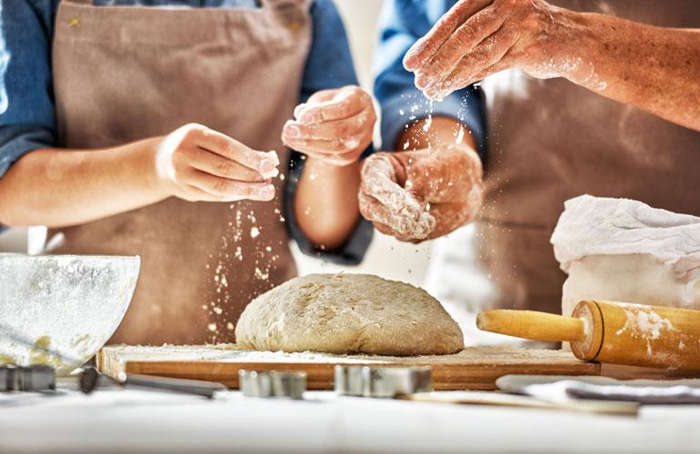 Man and Woman in a kitchen sprinkling flour over dough on a butcher block.